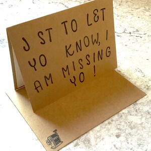 Puede incluir: Tarjeta marr&oacute;n plegada con el mensaje escrito a mano: "JST TO L&T YO KNOW, I AM MISSING YO!". La tarjeta tiene una peque&ntilde;a ilustraci&oacute;n de un sacapuntas. La tarjeta est&aacute; abierta, revelando un interior blanco.
