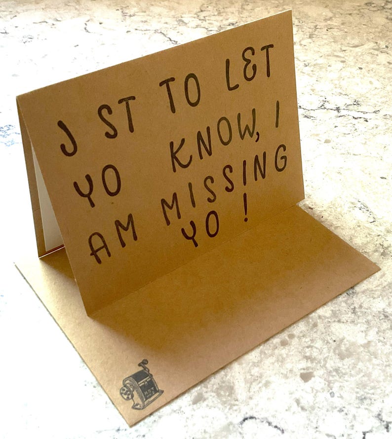 Puede incluir: Tarjeta marr&oacute;n plegada con el mensaje escrito a mano: "JST TO L&T YO KNOW, I AM MISSING YO!". La tarjeta tiene una peque&ntilde;a ilustraci&oacute;n de un sacapuntas. La tarjeta est&aacute; abierta, revelando un interior blanco.