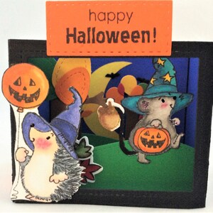 May include: A Halloween card with a pop-up scene featuring a hedgehog and a mouse dressed as witches. The hedgehog is holding a pumpkin balloon and the mouse is carrying a pumpkin. The card has a black frame and an orange banner that says "happy Halloween!"