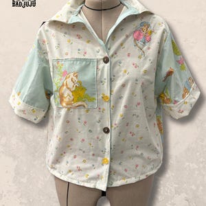 Puede incluir: Camisa reciclada con botones, estampado floral y detalles en azul claro. Presenta un dibujo de gato en el bolsillo y las mangas. La camisa tiene mangas cortas y está abotonada. El logotipo de la marca "UPCYCLED BADJUJU" es visible arriba a la izquierda.