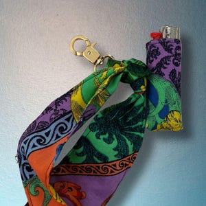 May include: A decorative lighter cover with a colorful scarf-like strap and a gold-colored clasp. The lighter cover is purple with a floral design. The strap features orange, green, purple, and blue patterns.