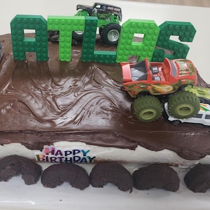Puede incluir: Una tarta de cumpleaños decorada con glaseado de chocolate, camiones monstruo de juguete y el nombre "ATLAS" escrito con bloques de construcción verdes. También se ve una vela con el número 4 y las palabras "FELIZ CUMPLEAÑOS".