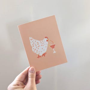 Puede incluir: Una tarjeta de felicitación con fondo melocotón que presenta una gallina blanca con manchas marrones y una cresta roja. La gallina está junto a un cóctel rojo con pajita y rodaja de limón. La tarjeta está sujeta por una mano.