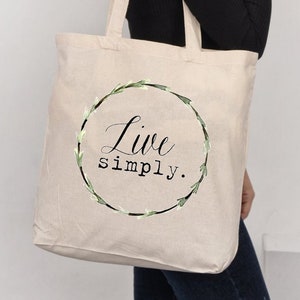 以下が含まれることがあります： 緑色の花の輪のデザインと「Live simply.」という黒い文字が印刷されたベージュのキャンバス地のトートバッグです。