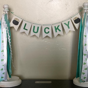 Puede incluir: Una pancarta blanca con la palabra "LUCKY" en letras verdes, decorada con amuletos de olla de oro. La pancarta está colgada entre dos soportes blancos con cintas de color verde, blanco y dorado.