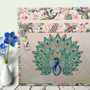 May include: A quilted beige zippered pouch featuring an embroidered peacock with a fanned tail in shades of green and blue. The top border and zipper pull are decorated with a colourful floral and peacock pattern on a cream background.