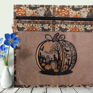 May include: A rectangular zippered pouch featuring a brown fabric body with a black embroidered pumpkin design containing a graveyard scene with tombstones, trees, and bats. The top border has a repeating pattern of pumpkins and ghosts.
