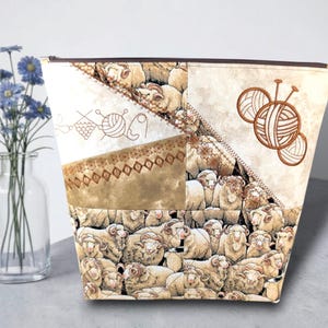 May include: A beige and brown fabric knitting project bag with a sheep pattern. The bag features embroidered yarn balls and knitting needles, and a zip closure. The bag is designed to hold knitting supplies.