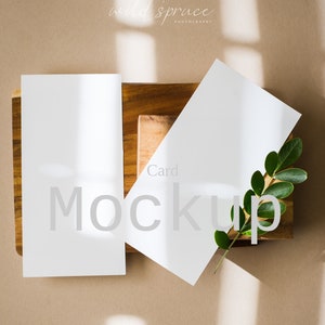 May include: Two blank white cards on a wooden surface with a green leaf branch. The text "Card Mockup" is visible on the cards. The background is a light brown colour with natural light shining through.