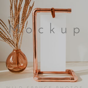 May include: A copper pipe stand with a blank white card mockup. The stand has a leather strap and is sitting on a white surface. A vase with dried brown grass is in the background.