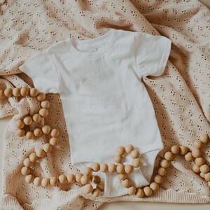 May include: A white baby onesie on a beige crocheted blanket with a wooden bead garland.
