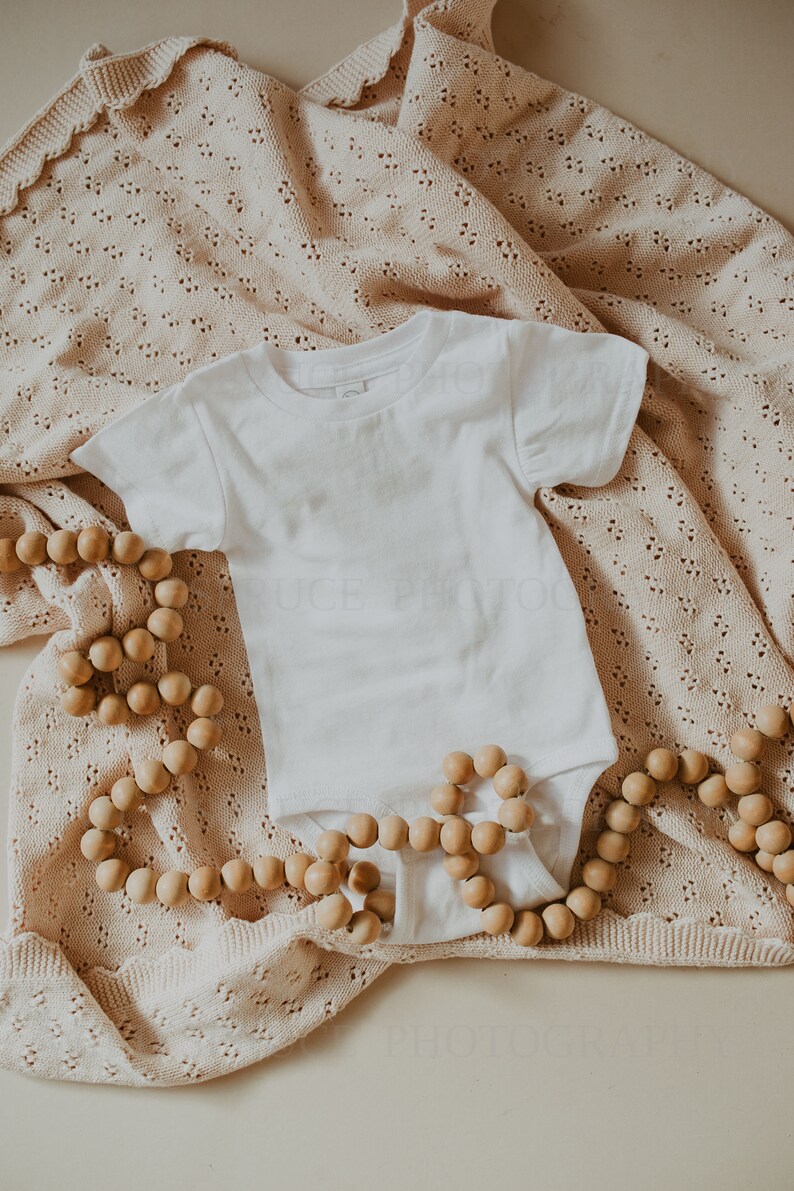 May include: A white baby onesie on a beige crocheted blanket with a wooden bead garland.