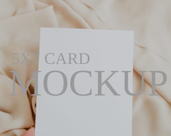 May include: A blank white rectangular card with the text 5X7 CARD MOCKUP printed in grey across the centre. The card is held against a soft, draped, light beige fabric background, providing a clean and neutral surface for design display.