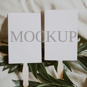May include: Two blank white cards with the word "MOCKUP" printed on them are attached to wooden sticks and placed on a beige fabric background with green leaves.