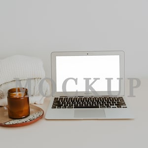 Può includere: Un laptop argentato con uno schermo bianco vuoto è su una scrivania bianca, accanto a una candela accesa in un barattolo di vetro marrone e una pila di coperte bianche. La parola "MOCKUP" è visibile sullo schermo. L'immagine è contrassegnata con "wild spruce photography".
