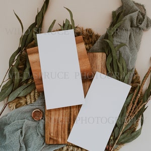 K&ouml;nnte beinhalten: Zwei leere wei&szlig;e Karten auf einem Holzbrett mit einer gewebten Basis, umgeben von gr&uuml;nem Laub und einem blauen Stoff.