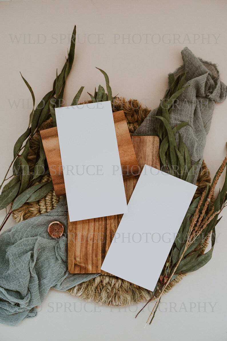 K&ouml;nnte beinhalten: Zwei leere wei&szlig;e Karten auf einem Holzbrett mit einer gewebten Basis, umgeben von gr&uuml;nem Laub und einem blauen Stoff.