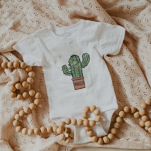 May include: A white baby onesie with a green cactus illustration in a terracotta pot. The cactus has yellow lights strung around it. The onesie is lying on a beige crocheted blanket with a wooden bead garland.