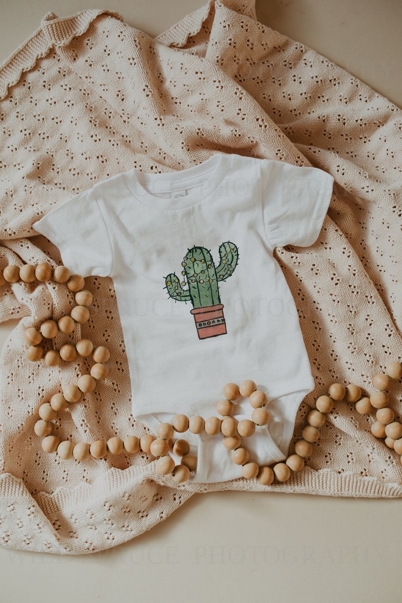 May include: A white baby onesie with a green cactus illustration in a terracotta pot. The cactus has yellow lights strung around it. The onesie is lying on a beige crocheted blanket with a wooden bead garland.