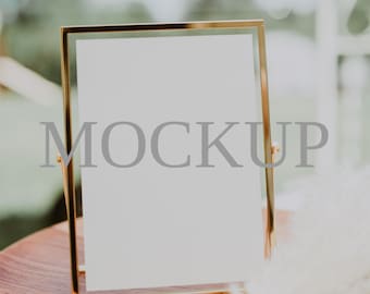 May include: A gold-colored picture frame with a white background and the word "MOCKUP" printed on it. The frame is sitting on a wooden table with a white fluffy feather plume in the foreground.