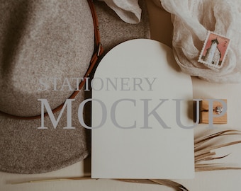 Può includere: Un biglietto bianco con il testo "STATIONERY MOCKUP" su uno sfondo beige. Il biglietto è circondato da un cappello marrone, un pezzo di stoffa bianca ed erba secca.