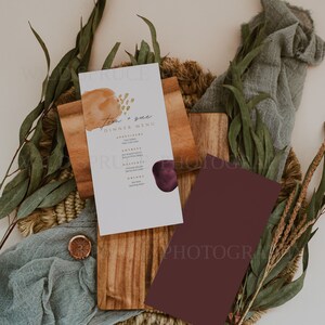K&ouml;nnte beinhalten: Eine Hochzeitsmen&uuml;karte mit einem Aquarellhintergrund in Braun- und Lilat&ouml;nen. Die Karte tr&auml;gt den Titel "Tom & Sue Dinner Menu" und listet Vorspeisen, Hauptgerichte, Desserts und Getr&auml;nke auf. Die Karte liegt auf einer Holzoberfl&auml;che mit einer geflochtenen Matte und gr&uuml;nem Laub.