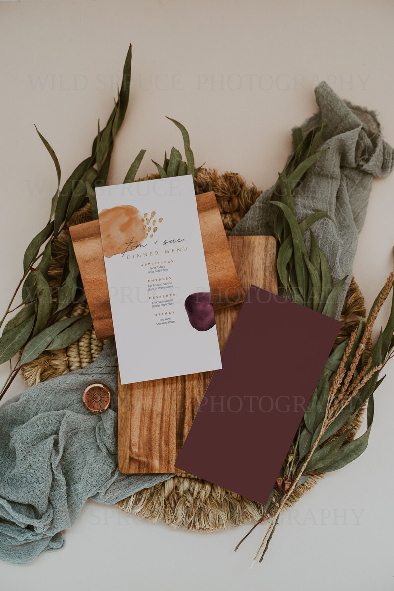 K&ouml;nnte beinhalten: Eine Hochzeitsmen&uuml;karte mit einem Aquarellhintergrund in Braun- und Lilat&ouml;nen. Die Karte tr&auml;gt den Titel "Tom & Sue Dinner Menu" und listet Vorspeisen, Hauptgerichte, Desserts und Getr&auml;nke auf. Die Karte liegt auf einer Holzoberfl&auml;che mit einer geflochtenen Matte und gr&uuml;nem Laub.