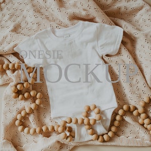 May include: White onesie mockup on a beige crocheted blanket with a wooden bead garland. The onesie has a crew neck and short sleeves. The text "ONESIE MOCKUP" is visible on the onesie.