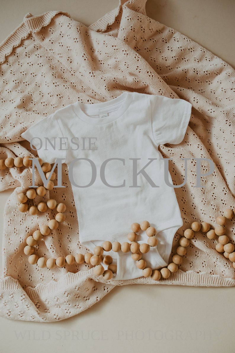May include: White onesie mockup on a beige crocheted blanket with a wooden bead garland. The onesie has a crew neck and short sleeves. The text "ONESIE MOCKUP" is visible on the onesie.