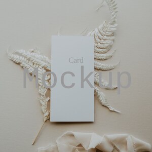 May include: A white card mockup with the word "Card" printed on it. The card is surrounded by dried white ferns and a beige ribbon with the text "wild spruce photography" printed on it.