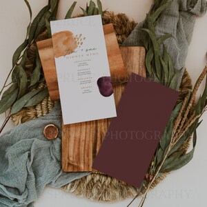 K&ouml;nnte beinhalten: Eine wei&szlig;e Karte mit einem braunen und goldenen Aquarelldesign und dem Text "Tom & Sue Dinner Menu" darauf gedruckt. Die Karte liegt auf einem Holzbrett mit einer burgunderfarbenen Karte daneben. Das Brett liegt auf einer gewebten Matte mit gr&uuml;nen Bl&auml;ttern und einem St&uuml;ck blauen Stoffes.