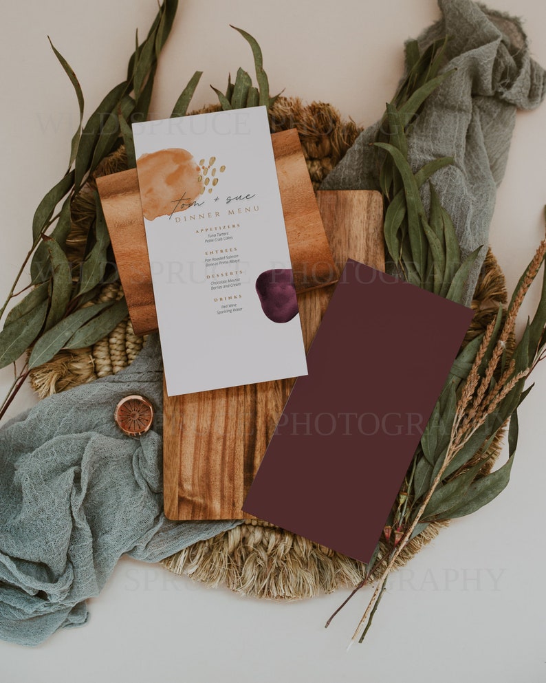 K&ouml;nnte beinhalten: Eine wei&szlig;e Karte mit einem braunen und goldenen Aquarelldesign und dem Text "Tom & Sue Dinner Menu" darauf gedruckt. Die Karte liegt auf einem Holzbrett mit einer burgunderfarbenen Karte daneben. Das Brett liegt auf einer gewebten Matte mit gr&uuml;nen Bl&auml;ttern und einem St&uuml;ck blauen Stoffes.