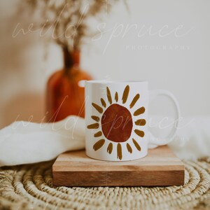 May include: White ceramic mug with a brown and gold sun design. The mug is sitting on a wooden coaster and a woven placemat.