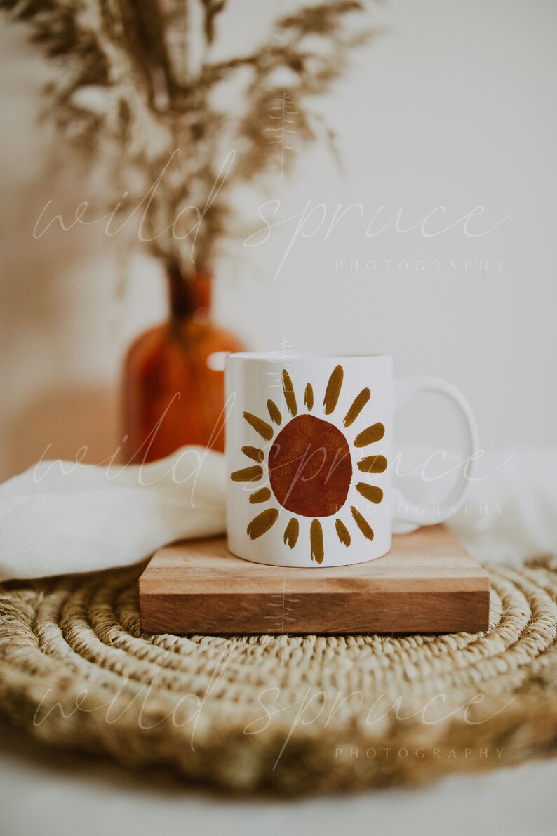 May include: White ceramic mug with a brown and gold sun design. The mug is sitting on a wooden coaster and a woven placemat.