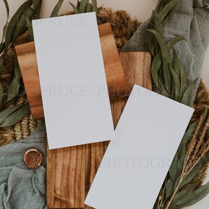 K&ouml;nnte beinhalten: Zwei leere wei&szlig;e Karten auf einer Holzoberfl&auml;che mit einer gewebten Basis und gr&uuml;nen Eukalyptusbl&auml;ttern. Die Karten sind perfekt, um Ihren eigenen Text oder Ihre eigenen Bilder hinzuzuf&uuml;gen.