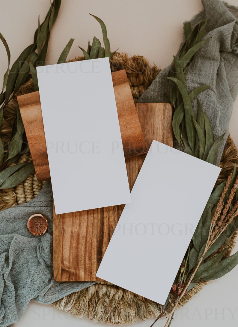K&ouml;nnte beinhalten: Zwei leere wei&szlig;e Karten auf einer Holzoberfl&auml;che mit einer gewebten Basis und gr&uuml;nen Eukalyptusbl&auml;ttern. Die Karten sind perfekt, um Ihren eigenen Text oder Ihre eigenen Bilder hinzuzuf&uuml;gen.