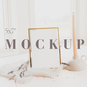Pode incluir: Uma moldura dourada de 5x7 polegadas está sobre uma superfície branca, ao lado de um castiçal branco com uma vela pêssego acesa. Lavanda seca e um tecido branco contribuem para a estética minimalista.