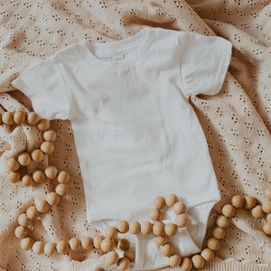 May include: A white baby onesie on a beige crocheted blanket with a wooden bead garland.