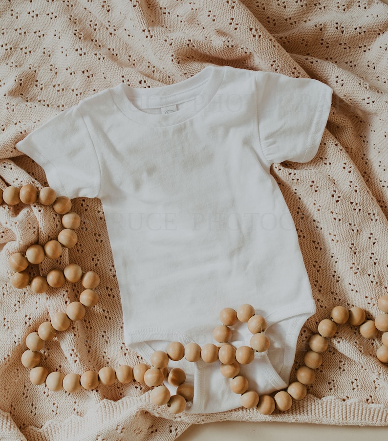 May include: A white baby onesie on a beige crocheted blanket with a wooden bead garland.