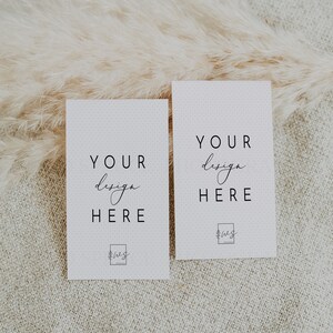May include: Two white cards with the text "YOUR design HERE" and a small logo at the bottom. The cards are on a beige textured surface with a fluffy white pampas grass behind them.