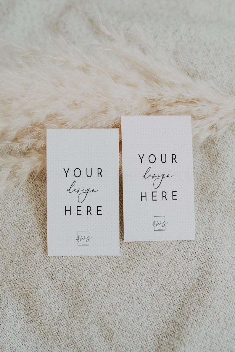 May include: Two white cards with the text "YOUR design HERE" and a small logo at the bottom. The cards are on a beige textured surface with a fluffy white pampas grass behind them.