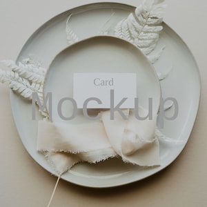 Puede incluir: Una tarjeta blanca con la palabra "Card" impresa en ella, colocada en un plato blanco con una cinta de tela blanca y helechos blancos secos. El plato está sobre una superficie beige. El texto "Mockup" está superpuesto a la imagen.