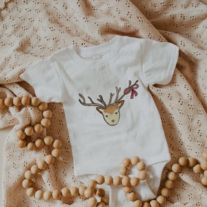 May include: A white baby onesie with a brown and white reindeer illustration and a red bow. The onesie is lying on a beige knitted blanket with wooden beads.