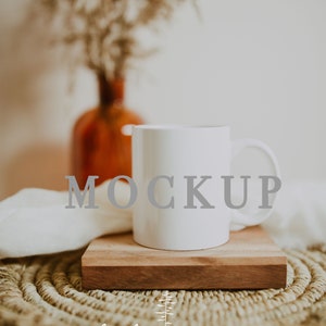 May include: White ceramic mug with the word "MOCKUP" printed on it, sitting on a wooden coaster on a woven straw mat. The mug is in focus, and the background is blurred.