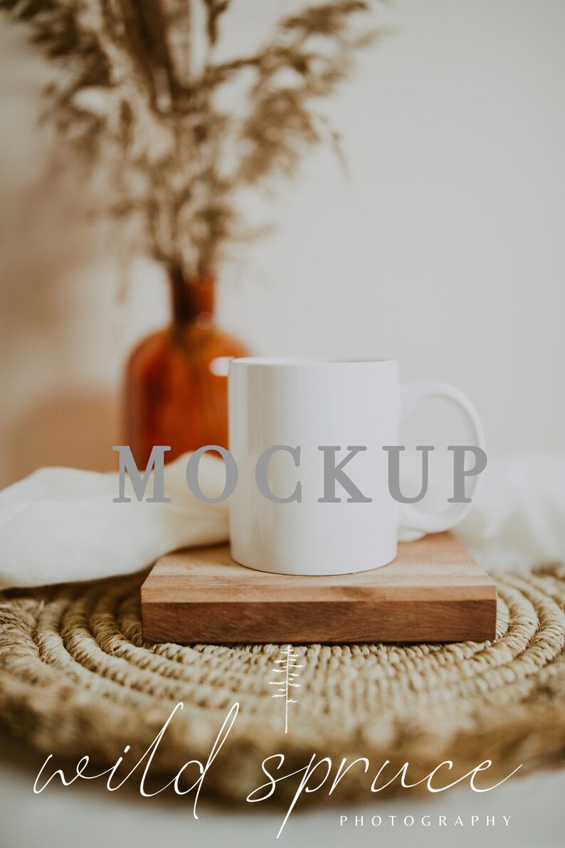 May include: White ceramic mug with the word "MOCKUP" printed on it, sitting on a wooden coaster on a woven straw mat. The mug is in focus, and the background is blurred.