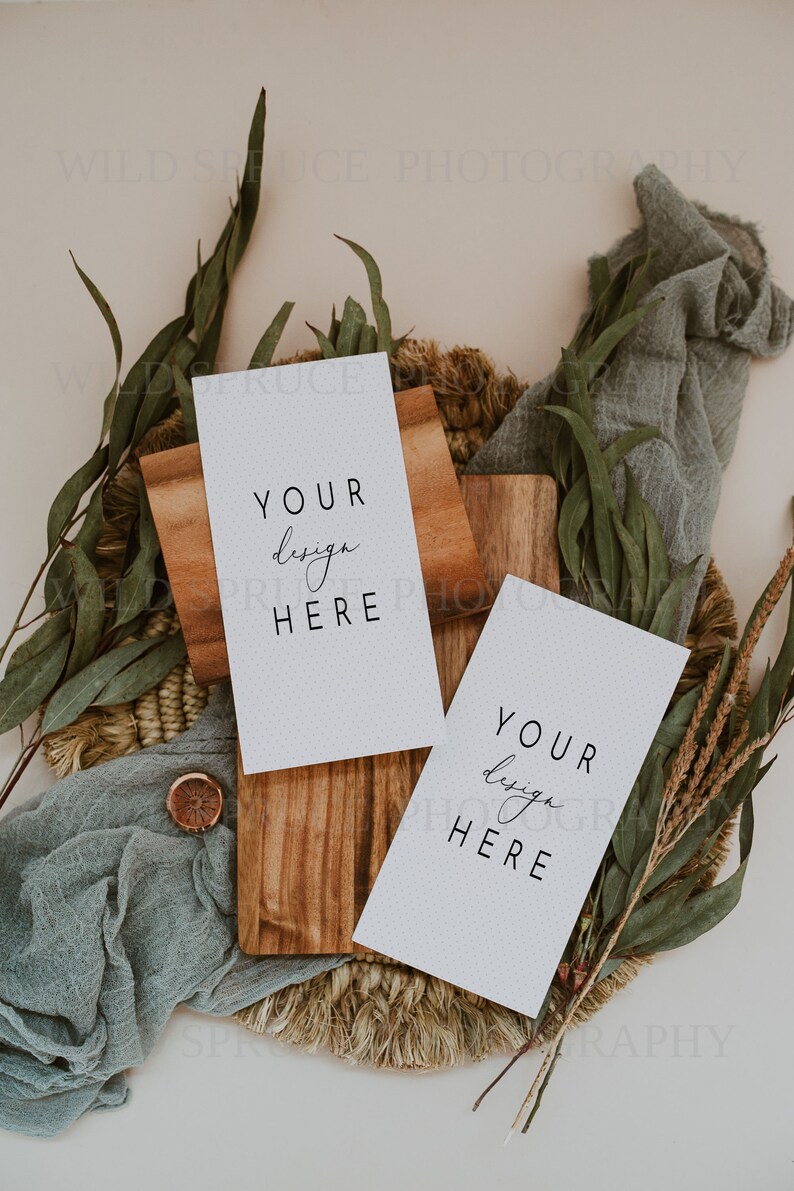 K&ouml;nnte beinhalten: Zwei leere wei&szlig;e Karten mit dem Text "Your design here" auf einer Holzoberfl&auml;che mit einer geflochtenen Matte und Gr&uuml;npflanzen.