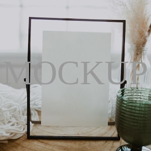 以下が含まれることがあります： 前面に「MOCKUP」の文字が印刷された透明なガラスフレームが、木製の表面に置かれています。緑色のガラスのゴブレットと、ドライフラワーが入った花瓶も確認できます。明るい部屋で、ディスプレイやプレゼンテーションのシーンを演出しています。