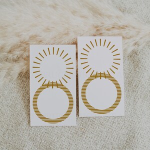 May include: Two white cards with gold sunburst and striped circle designs. The cards are on a beige textured surface.