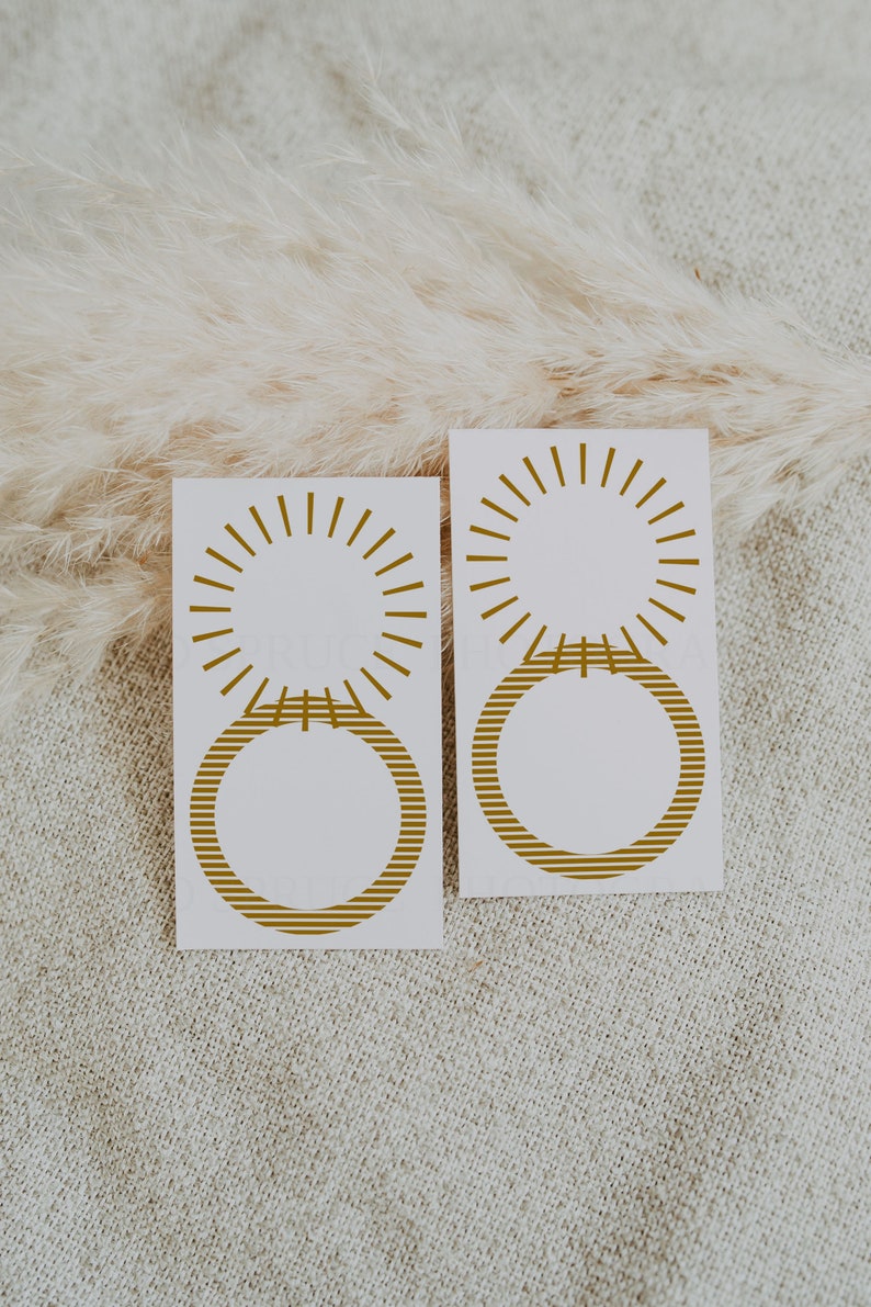 May include: Two white cards with gold sunburst and striped circle designs. The cards are on a beige textured surface.