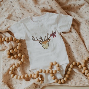 May include: A white baby onesie with a brown and red reindeer illustration. The onesie is lying on a beige knitted blanket with wooden beads.